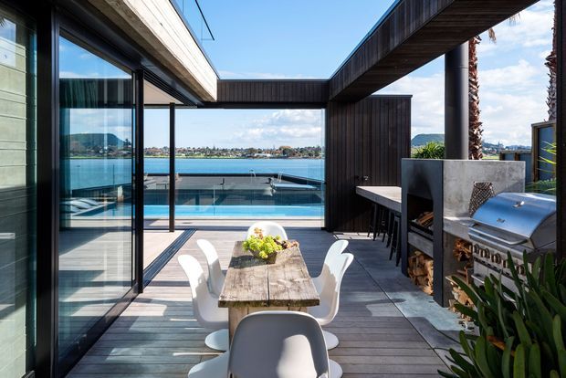 A contemporary waterfront outdoor dining terrace with a timber dining table and white chairs, overlooking a swimming pool and bay. Dark timber-clad walls, large glazed sliding doors, exposed structural beams, and a built-in stainless-steel barbecue define the space.