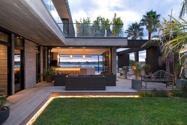 A sophisticated contemporary outdoor living area arranged around a small grass courtyard, with dark timber or wood-look cladding, concrete and timber-toned surfaces, built-in seating, dining furniture, planting, and warm linear lighting. The open-plan terrace frames a visible waterfront beyond, while an upper-level balcony with glass balustrades overlooks the space.