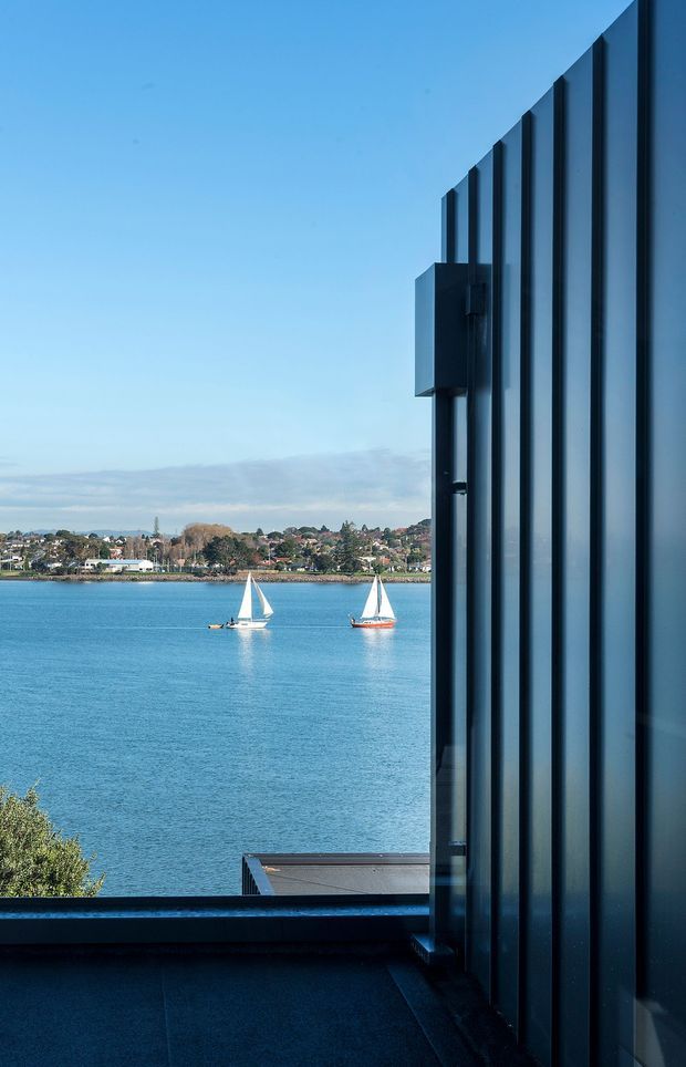 A contemporary waterfront architectural detail viewed from a dark terrace or balcony. The right side is dominated by tall vertical dark metal cladding or fins, while the open view looks across bright blue water toward a suburban shoreline with two sailboats. A dark deck or roof edge occupies the foreground, creating a strong contrast between the shadowed structure and the sunny coastal outlook.