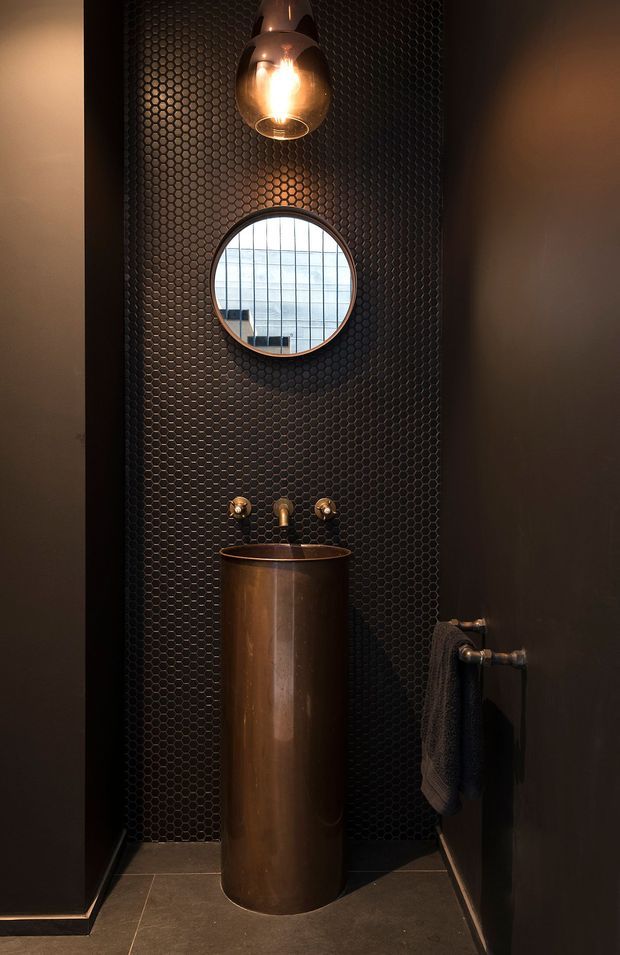 A narrow, moody bathroom featuring a cylindrical copper pedestal basin with exposed wall-mounted taps, dark hexagonal mosaic tilework, a round mirror, and a warm pendant light. Dark painted walls and dark floor tiles create a dramatic industrial-inspired setting, with a copper towel rail adding a metallic accent.