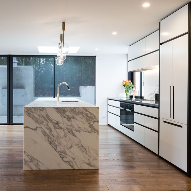 Contemporary kitchen featuring a large veined stone island with an integrated sink and slim black tap, white flat-panel cabinetry with black framing and handles, a built-in oven, and a reflective backsplash. Warm timber flooring contrasts with the bright white ceiling and cabinetry. Full-height dark-framed glazing opens toward an outdoor area, while pendant globes and recessed downlights provide illumination.