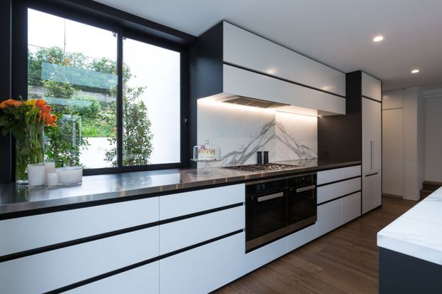 Contemporary kitchen with streamlined white flat-panel cabinetry, dark framing and accents, a marble-look splashback, dark benchtops, integrated appliances, timber flooring, and a large black-framed window overlooking greenery. Under-cabinet lighting adds a refined, functional feel.
