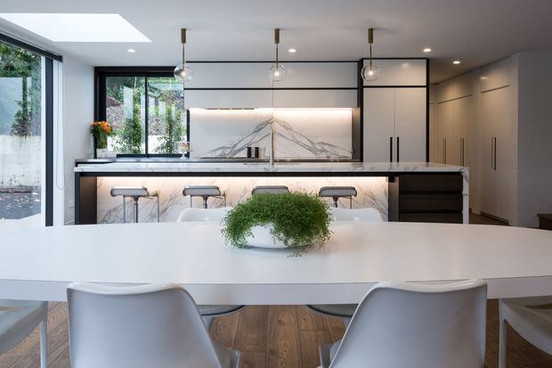 A bright, refined open-plan kitchen and dining area. The contemporary kitchen features a long island with waterfall-style stone surfaces, veined marble-look splashback, dark lower cabinetry, pale upper cabinetry, integrated appliances, pendant lights, and bar stools. A white dining table with modern curved chairs and a central green plant occupies the foreground. Large glazing and a skylight bring in abundant natural light.