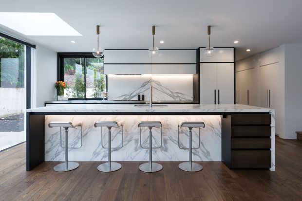 Contemporary kitchen with a large marble-look island and integrated breakfast bar, four bar stools, sleek dark-framed glass and cabinetry, pendant lighting, and timber flooring. Large glazing and a skylight bring in abundant natural light.
