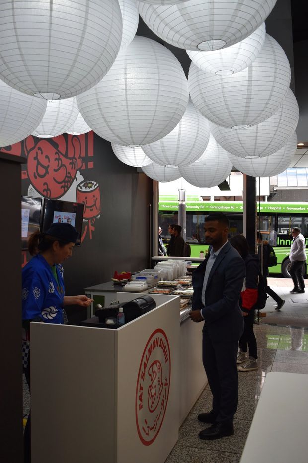 A compact Asian restaurant or takeaway counter inside a commercial space, with a white service counter, food trays, cashier, and customers. Numerous large white ribbed paper lanterns hang densely from the ceiling. A dark wall features a bold red graphic mural and restaurant branding, while glazing at the front looks onto a city street and passing bus.