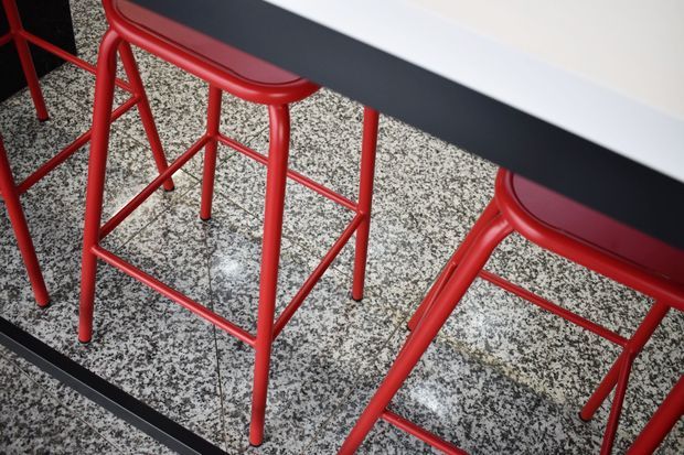 Close-up view of red metal-framed bar stools with dark seats positioned along a dark countertop or bar. The stools stand on a speckled terrazzo-like tiled floor, creating a bold red, black, and neutral palette. The image focuses on furniture and interior finishes rather than a complete room.