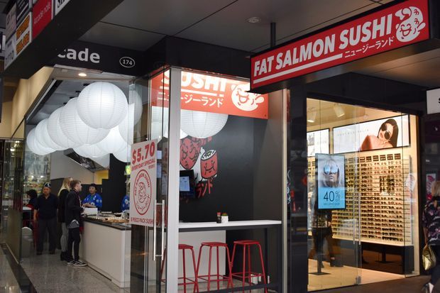 Street-level view of a compact Japanese sushi takeaway storefront in an indoor shopping arcade. The shop has bold red and black signage, illuminated paper lanterns, a narrow white counter with red stools, and visible food-service branding. A neighbouring retail storefront is visible through the glazing on the right.