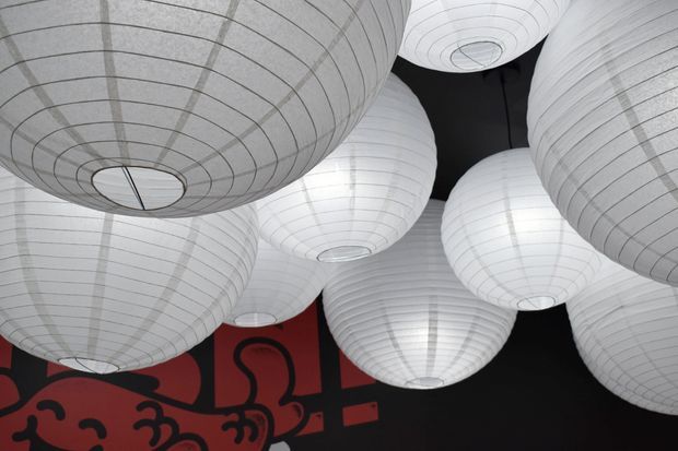 Close-up interior view of numerous oversized white ribbed paper lantern pendant lights clustered across a dark ceiling. A bold red-and-black graphic wall or mural is partially visible below, creating a dramatic contemporary installation.
