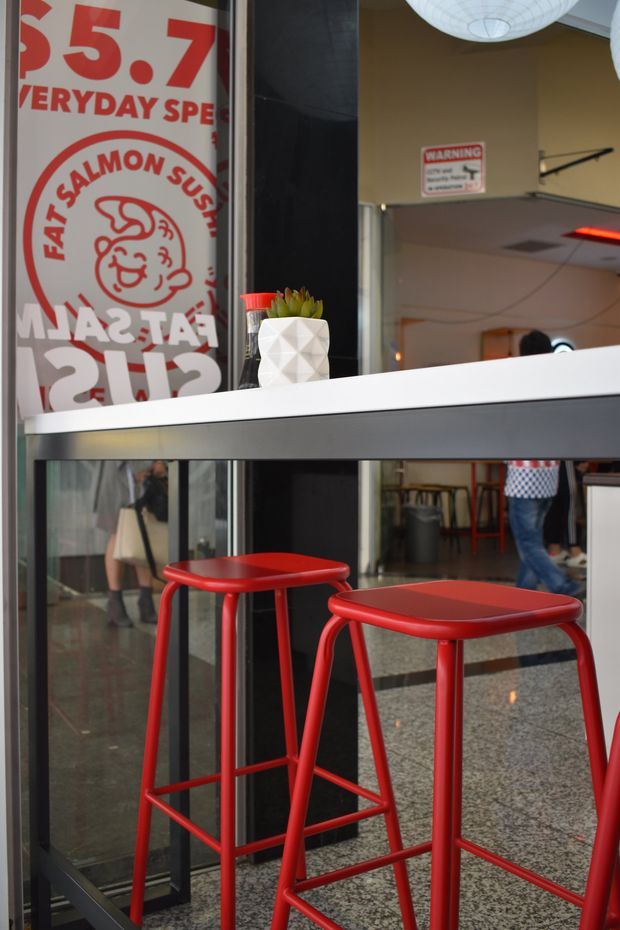A close-up view of a casual fast-food restaurant interior, showing a white and grey service counter with a small geometric white planter, red metal bar stools, dark-framed glass partitions, terrazzo flooring, and bold red-and-white promotional signage. The space has a playful, retro-commercial character with visible overhead pendant lighting and food-service details.