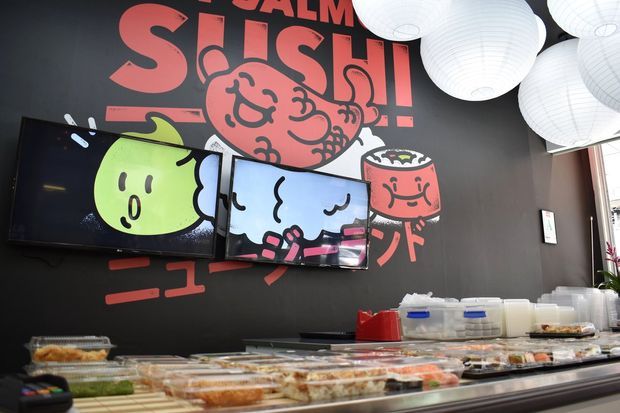 A vibrant sushi restaurant interior viewed from behind the sushi display counter. The dark feature wall is covered with large red and pink cartoon-style graphic artwork and text, with multiple wall-mounted screens integrated into the mural. White paper lantern pendant lights hang overhead, while trays of sushi and food containers fill the foreground counter.