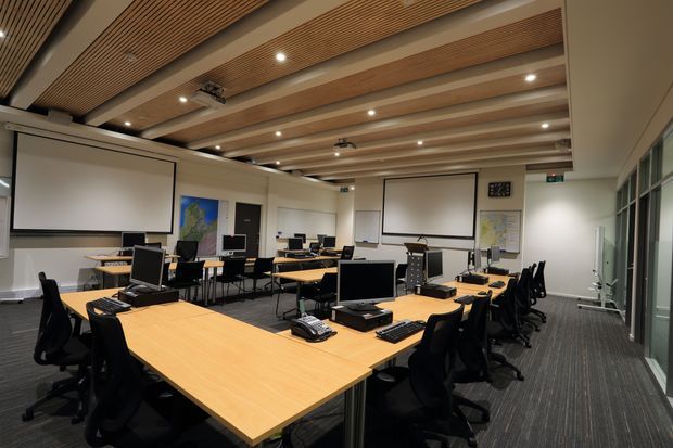 A modern computer training or teaching room with multiple grouped timber tables, black ergonomic office chairs, desktop computers, keyboards and telephones. Several ceiling-mounted projection screens and projectors support presentations. The room has a linear timber-slat and exposed-beam ceiling, recessed spotlights, carpeted flooring, white walls, and glazed partitions along one side.