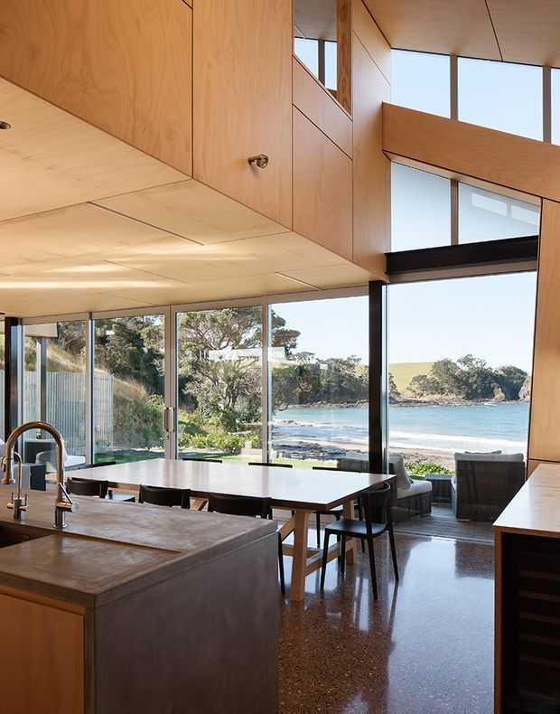 A contemporary open-plan kitchen and dining area with extensive floor-to-ceiling glazing overlooking a beach and sheltered coastal inlet. The room features warm timber cabinetry and ceiling panels, a large stone or composite island with integrated sink, a long dining table, dark-framed windows, polished concrete or terrazzo-like flooring, and a compact living area near the glazing. Natural light and the coastal outlook dominate the composition.