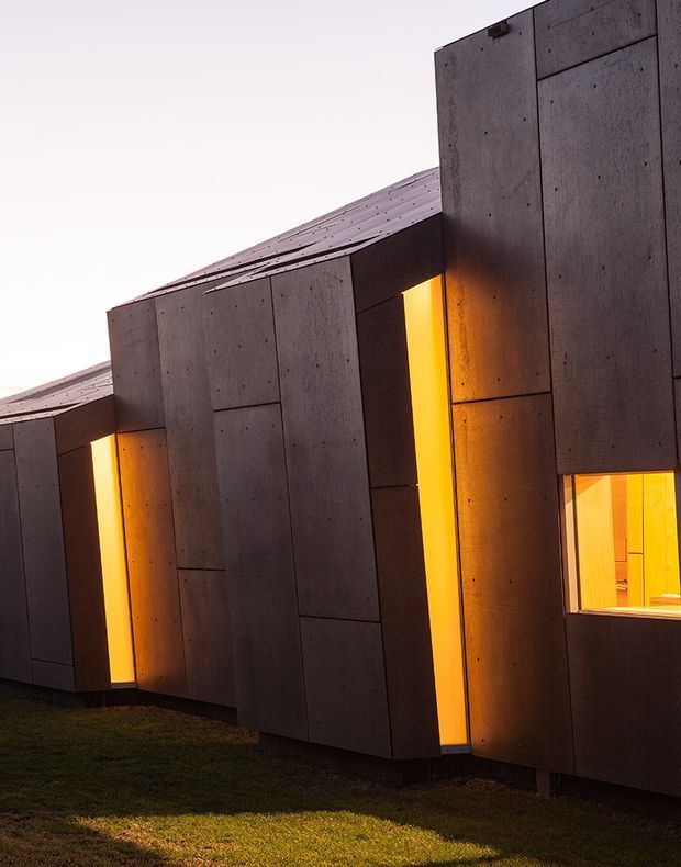 A dramatic close exterior view of a contemporary, angular building clad in dark grey concrete panels. Narrow vertical openings and a larger window emit warm amber light, creating strong contrast against the textured facade. The building has a sloping metal roof and sits beside a grass-covered site at dusk.