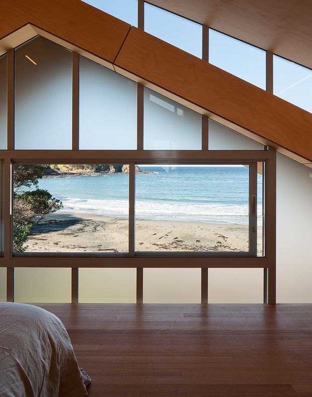 A serene bedroom interior with a bed partially visible in the foreground, warm timber flooring and ceiling, and extensive dark timber-framed glazing. The angled timber wall and clerestory windows create a distinctive architectural composition, with direct views to a sandy beach and turquoise sea beyond.