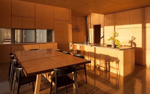 Warm, sunlit open-plan kitchen and dining area with extensive timber wall and ceiling panelling, integrated timber cabinetry, a large central island, and a substantial timber dining table with dark chairs. Strong late-afternoon sunlight enters through the glazing, creating a rich amber atmosphere and reflections across the polished floor.