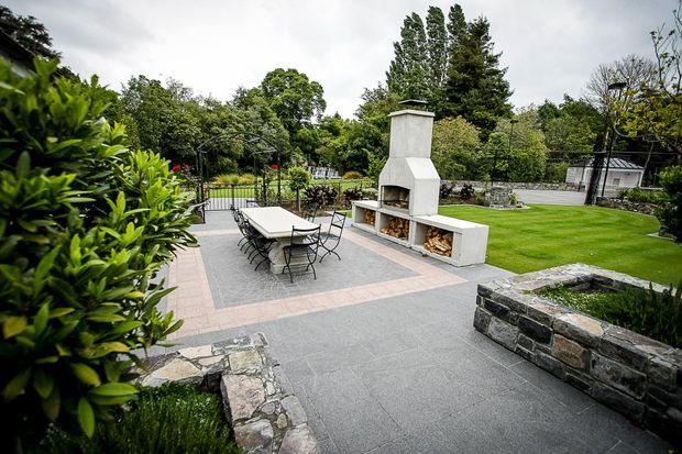 A landscaped outdoor entertaining area with a paved terrace, dining table and chairs, a built-in white masonry fireplace with timber storage, manicured lawn, established planting, and low natural-stone retaining walls. The setting appears spacious and garden-oriented, with a refined contemporary outdoor design.
