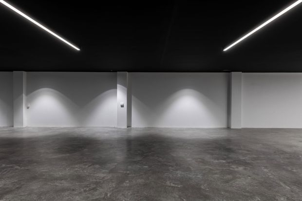 Empty, minimalist interior with a broad polished grey concrete floor, white structural walls and a black ceiling. Linear recessed or surface-mounted strip lights create strong symmetrical illumination across the walls, suggesting a gallery, showroom, warehouse-conversion, or other commercial space.