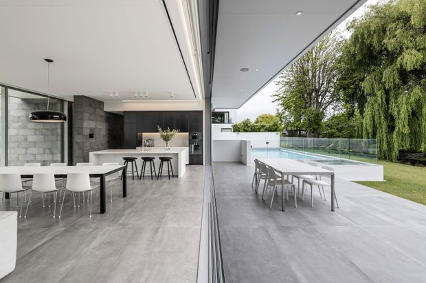 A contemporary open-plan kitchen and dining area extending seamlessly onto a covered outdoor terrace. The interior features a white island with stools, a dark timber kitchen wall, grey stone-look flooring, exposed concrete-block walls, and a dining table with white chairs. Full-height glazed sliding doors create a strong indoor–outdoor connection, with an outdoor dining setting and swimming pool visible beyond.