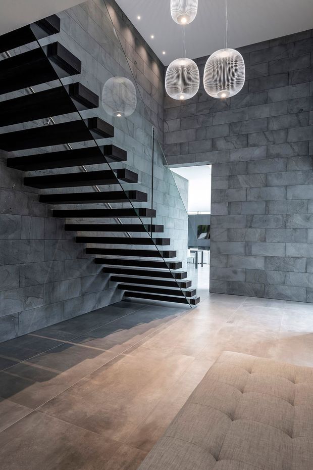 A dramatic contemporary interior featuring a floating black open-riser staircase with a glass balustrade, set against large-format dark grey stone or concrete-look wall tiles. Three sculptural pendant lights hang over the stair and entry zone. The space has a polished concrete-look floor, high ceiling, and a partial view of a tufted upholstered sofa in the foreground.