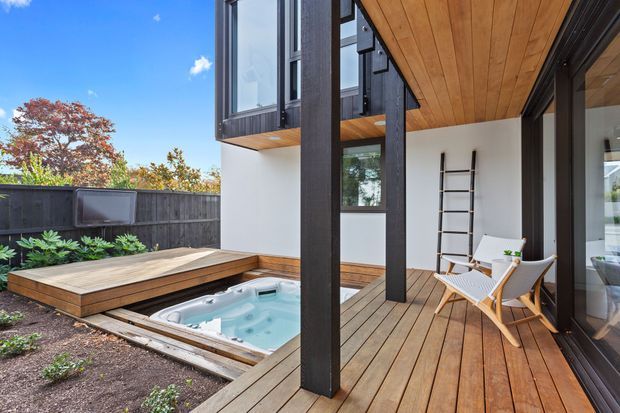A contemporary timber-decked outdoor living area beside a small built-in spa pool, framed by dark structural posts and large glazed doors. The covered deck includes light timber lounge chairs, a ladder-like wall feature, and a raised timber platform, with a dark boundary fence and landscaped planting beyond.