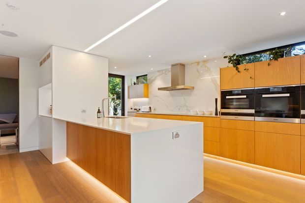 A bright contemporary kitchen featuring extensive warm timber cabinetry, a large white island with an integrated sink, white marble-look splashback, stainless-steel rangehood, and built-in black appliances. LED strip lighting beneath the cabinetry and island adds a refined glow, while timber flooring and indoor planting soften the space.