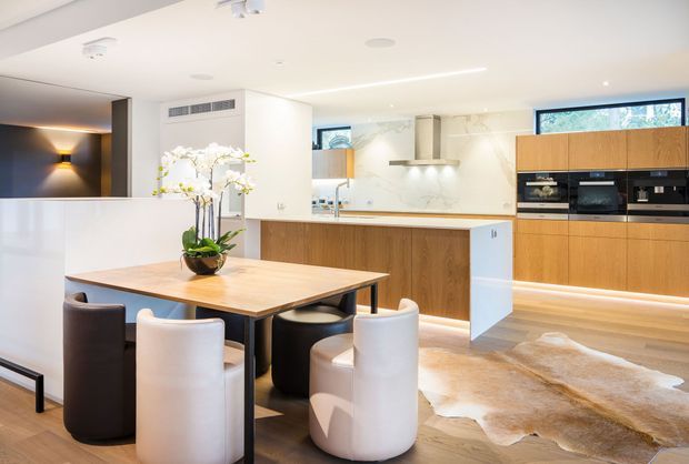 A bright, open-plan contemporary kitchen and dining area with a large timber dining table, mixed upholstered dining chairs, timber cabinetry, a white stone island and splashback, integrated appliances, and soft under-cabinet lighting. A partial living or media area is visible at left, while a natural hide rug lies over the timber flooring.
