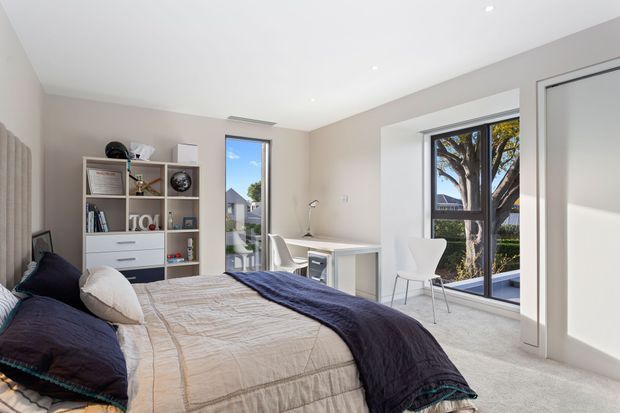Bright contemporary bedroom with a neatly made bed, neutral upholstered headboard, open shelving and drawers, a compact desk, and a white chair. Full-height glazed doors and a tall window provide abundant natural light and views of a tree and neighbouring homes. The room has soft grey-beige carpet, pale walls, and navy and neutral bedding.
