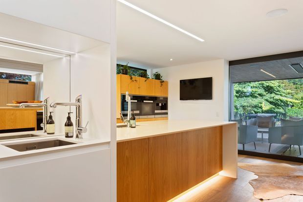 Bright contemporary open-plan kitchen with a central island clad in warm timber cabinetry and a pale benchtop, integrated sink and tap, and a compact cooking zone with mustard-yellow overhead cabinetry. The kitchen opens to a dining or outdoor entertaining area through large dark-framed glazing, with indoor greenery, recessed linear lighting, and a natural hide rug adding warmth.