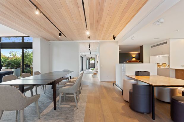 Open-plan contemporary dining and kitchen interior with a large timber dining table, upholstered dining chairs, a white kitchen island, dark curved bar stools, pale timber flooring, and a slatted timber ceiling with integrated track lighting. Full-height glazing brings in abundant daylight and opens toward a landscaped outdoor area.