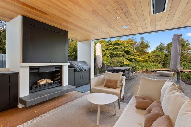 Covered outdoor entertaining area with a timber-lined ceiling, built-in fireplace, barbecue, dining setting and relaxed lounge seating opening onto a landscaped deck. The space has a refined contemporary resort-like feel, with warm timber finishes, neutral upholstery and dark built-in elements.