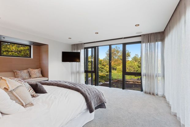 Bright contemporary bedroom with a large bed, soft grey carpet, full-height black-framed glazing and sheer curtains overlooking leafy greenery. A timber-panelled bedhead niche and wall-mounted television add built-in detailing.