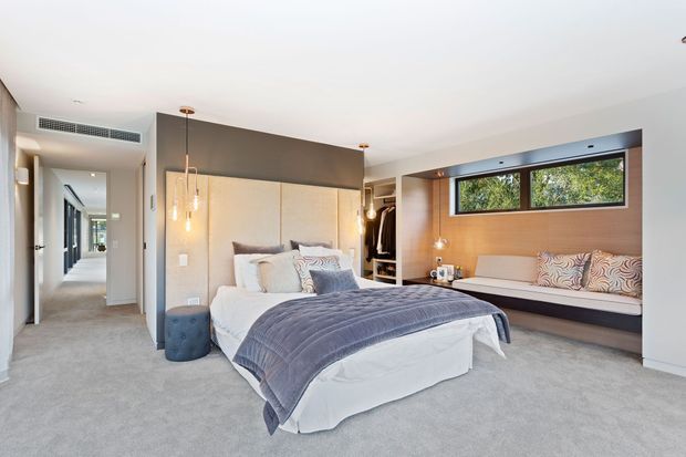 A bright contemporary bedroom with a large upholstered bed, layered white and grey bedding, and a soft carpeted floor. The room features a timber-lined window wall with a built-in upholstered window seat, dark grey feature walls, built-in wardrobe joinery, pendant lights, and a hallway visible beyond the open doorway.