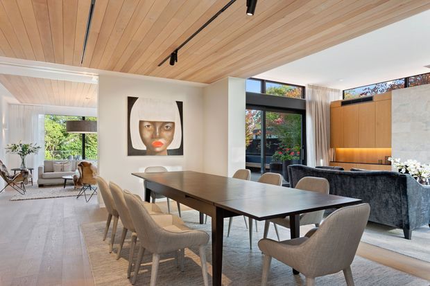 A refined open-plan dining and living area with a long dark timber dining table surrounded by upholstered chairs. The space features pale timber wall and ceiling lining, light flooring, large black-framed windows, a warm timber built-in cabinet, and a textured stone fireplace wall. Contemporary furnishings and a large artwork create a calm, natural, sophisticated interior.