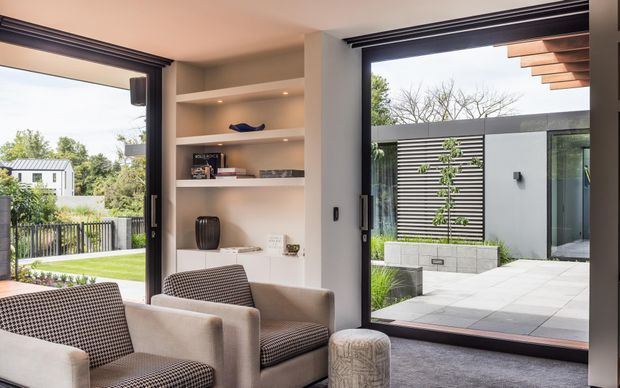 Contemporary living room with built-in recessed shelving, two upholstered armchairs, and a small ottoman, opening through large black-framed glazed doors to a landscaped courtyard and adjacent modern pavilion. The room has neutral plaster walls, dark trim, and a carpeted floor, with strong indoor-outdoor visual connection.