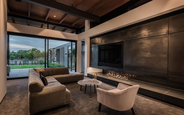 A sophisticated contemporary living room with a large sectional sofa, sculptural lounge chair, round coffee tables, and a dramatic built-in black feature wall incorporating a television and linear fireplace. Exposed timber ceiling boards and dark steel structural beams create an industrial character, while full-height glazing opens the room to a landscaped outdoor area.
