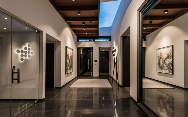 A sophisticated contemporary residential entry hall with glossy dark tiled flooring, white walls, black-framed full-height glazing and a large mirror reflecting the corridor. The space features a warm timber-lined ceiling with recessed spotlights, a dramatic skylit void, minimalist artwork, sculptural wall lighting and dark internal doors.