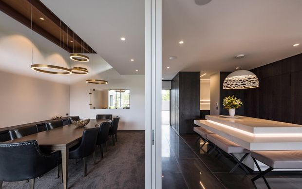 A refined contemporary dining and entertaining space viewed through a glass partition. On the left is a formal dining area with a dark rectangular table, black upholstered chairs, pendant lighting, and a wall mirror. On the right is a large kitchen island or dining counter with layered timber surfaces, integrated warm LED lighting, dark cabinetry, glossy dark flooring, and a sculptural pendant light. The palette is predominantly dark wood, black, warm white, and neutral grey.