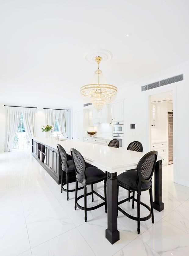 Bright, luxurious white kitchen with a large island and integrated dining bar, black bar stools, white cabinetry, glossy white flooring, and a prominent crystal chandelier with gold detailing. Tall curtained windows and ornate ceiling moulding contribute to a refined classic aesthetic.