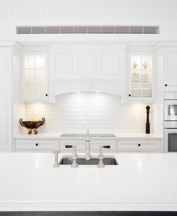 Bright, predominantly white traditional kitchen viewed from the island. The room features detailed panelled cabinetry, glass-front upper cupboards, a decorative rangehood surround, white subway-tile splashback, integrated cooktop, built-in appliances, and a large white island with a polished metal bridge faucet and undermount sink. Ornate ceiling cornice detailing adds a classic character.