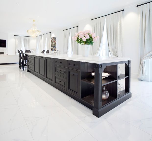 Bright, spacious open-plan kitchen and dining area dominated by a large dark charcoal island with paneled cabinetry, drawers, integrated open shelving, and a white stone countertop. The room features glossy white marble-look flooring, white walls and ceiling, tall windows with white drapery, a crystal chandelier, and restrained decorative styling with flowers and glassware.