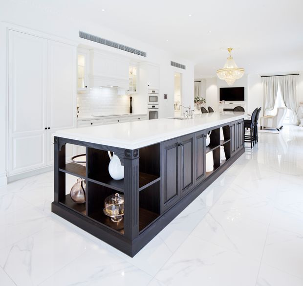 Bright, spacious white kitchen with a large dark timber island featuring paneled cabinetry and open display shelving. The island has a white stone benchtop and integrated seating, while the surrounding kitchen includes white paneled cabinetry, integrated appliances, a light tiled splashback, and polished white marble-look flooring. A crystal chandelier and adjoining dining/living area add a refined, classic feel.