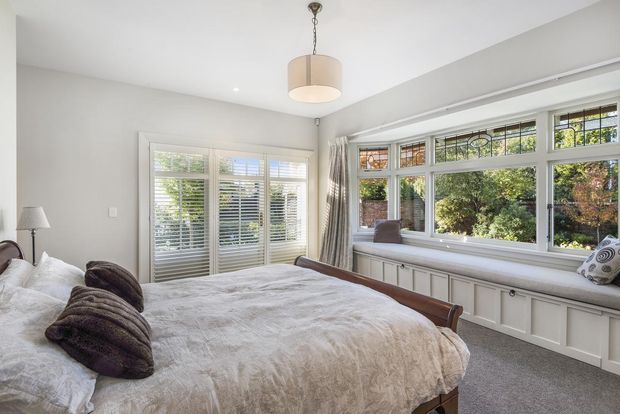 Bright, elegant bedroom with a large upholstered bed, soft neutral bedding, plantation shutters, and a built-in window seat beneath a wide bank of windows. The room has pale walls, grey carpeting, white paneled joinery, layered curtains, and a simple pendant light, creating a calm and refined atmosphere.