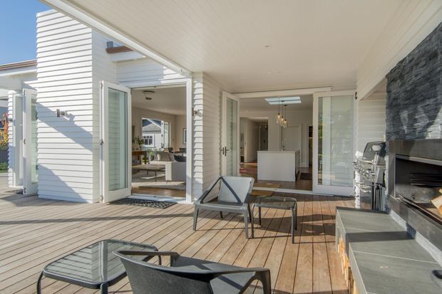 A bright covered outdoor deck or terrace adjoining a modern coastal-style home. The timber decking extends across the foreground with contemporary outdoor seating, while wide bi-fold or hinged glazed doors open into an interior living and kitchen area. White weatherboard walls, a pale-lined ceiling, large glazing, and a dark stacked-stone outdoor fireplace define the space.