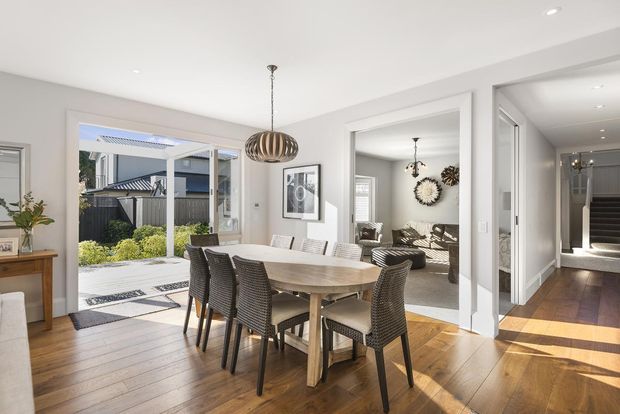 Bright open-plan dining area with a large timber dining table and woven dark dining chairs, connected to a living room and hallway. White walls and trim, warm timber flooring, pendant lighting, and expansive glazed doors opening to a landscaped outdoor area create an airy contemporary interior.