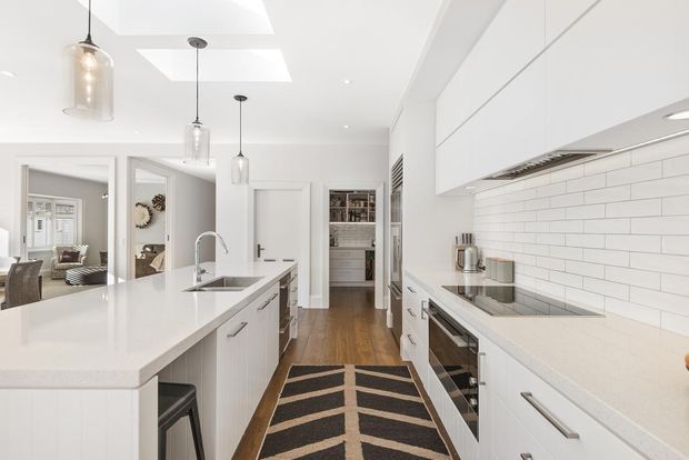 Bright, spacious contemporary white kitchen with a large central island, integrated sink and seating, extensive white cabinetry, stainless-steel appliances, and a white horizontal subway-tile splashback. Timber flooring, pendant lights, skylights, and a patterned runner add warmth and contrast. A living or dining area is visible through the open doorway.