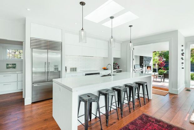 Bright contemporary white kitchen with a large central island, five dark metal stools, stainless-steel double-door refrigerator, white subway-tile splashback, extensive white cabinetry, pendant lights, skylights, and warm timber flooring. The kitchen opens toward a furnished outdoor terrace or courtyard.