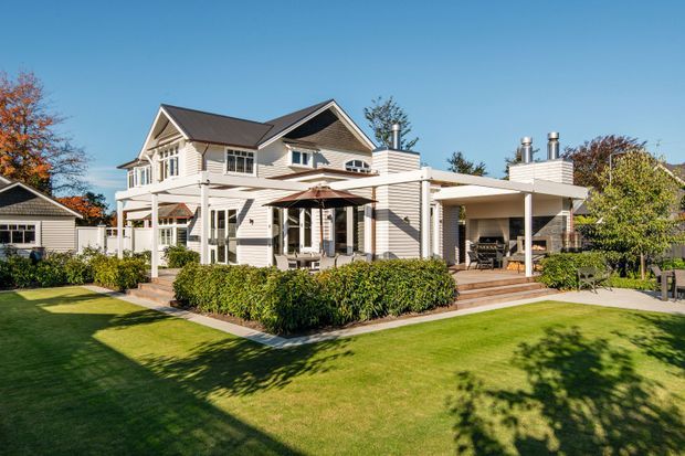 Exterior view of a large, light-clad suburban house with multiple pitched gables, white-framed windows, covered verandas, a timber deck, and an attached covered outdoor entertaining area. The landscaped garden includes manicured lawn and hedges, with a detached outbuilding visible to the left.