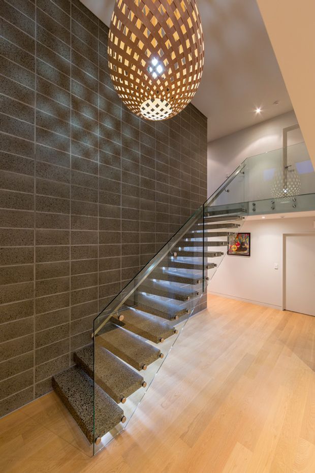 Interior view of a contemporary stairway and landing with open-riser stone or concrete steps, transparent glass balustrades, a dark textured tiled feature wall, warm timber flooring, and a large sculptural woven pendant light.