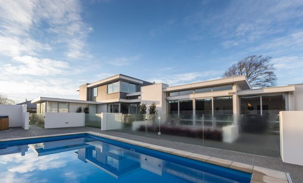 Exterior view of a large contemporary residential home arranged around a swimming pool. The building has clean rectangular volumes, flat rooflines, extensive glazing, pale rendered walls, dark window framing, and a covered outdoor entertaining area with glass balustrades. The pool and paved terrace dominate the foreground.