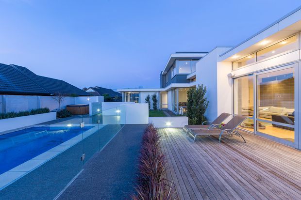 Contemporary residential pool area viewed from a timber deck at dusk. The scene features a long rectangular swimming pool with frameless glass fencing, integrated garden lighting, timber decking, dark gravel landscaping, and a modern white multi-level house with extensive glazed doors and windows. Sun loungers sit beside the pool, and a warmly lit interior is visible through the glazing.
